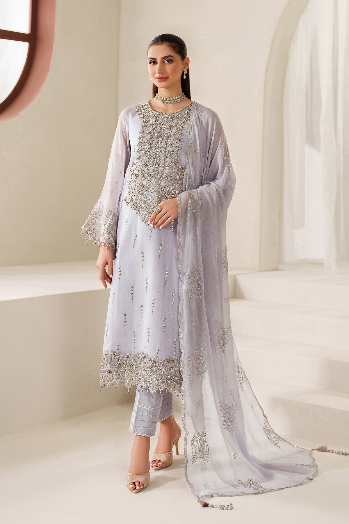 Woman wearing a light gray traditional outfit with silver embroidery in a bright room.